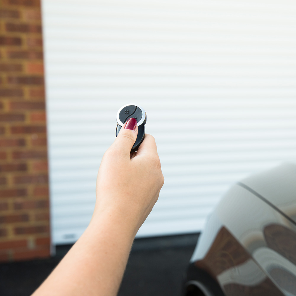 Close up of a hand holding out a SWS UK electric operation key fob with a white roller garage door visible in the background